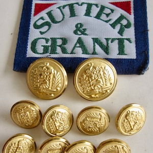 Puede incluir: Diez botones de color dorado con un diseño en relieve que presenta un escudo y el texto "Sutter & Grant". Los botones están dispuestos en semicírculo sobre una superficie blanca. Un parche de tela azul con el texto "Sutter & Grant" en letras blancas y verdes es visible en el fondo.