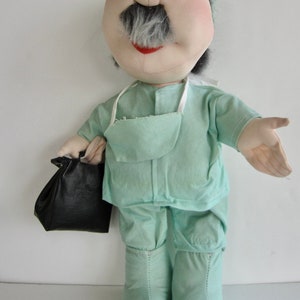 May include: A plush doctor doll wearing a green surgical scrub suit and a blue surgical cap. The doll has a white mustache and a black medical bag.