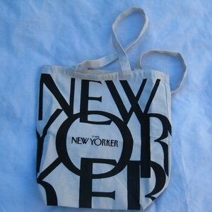 May include: A cream-colored tote bag with black text that reads "NEW YORKER". The bag has two cream-colored handles. The text "THE NEW YORKER" is printed in a smaller font size below the word "NEW".