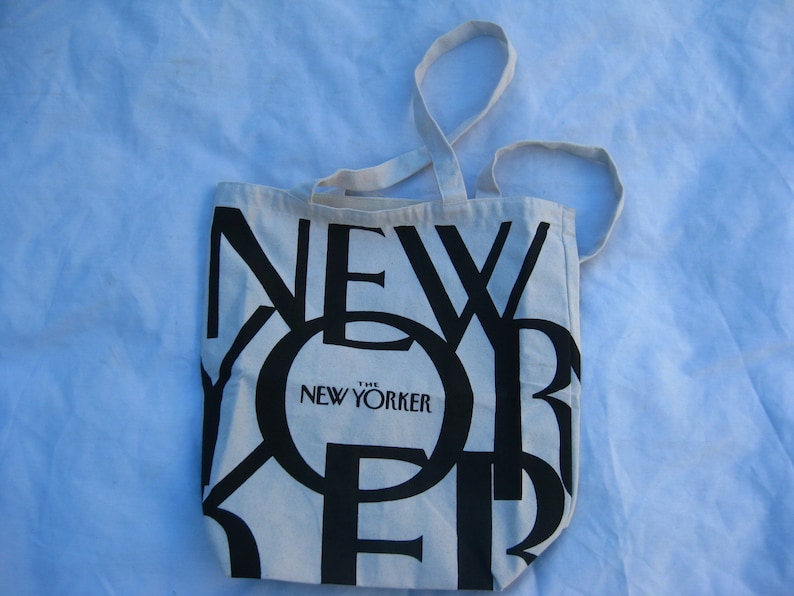 May include: A cream-colored tote bag with black text that reads "NEW YORKER". The bag has two cream-colored handles. The text "THE NEW YORKER" is printed in a smaller font size below the word "NEW".