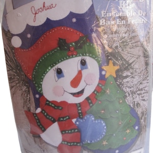 May include: A felt stocking kit featuring a snowman holding a blue mitten and a green Christmas tree. The kit includes cotton floss, needle, pattern, and instructions. The stocking is personalized with the name "Joshua".