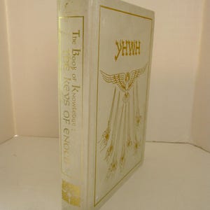 Peut inclure: Un livre &agrave; couverture rigide de couleur cr&egrave;me avec des lettres dor&eacute;es. Le titre "The Book of Knowledge: The Keys of Enoch" est imprim&eacute; sur le dos. La couverture avant pr&eacute;sente le mot "YHWH" et une illustration d'aigle avec des rayons.