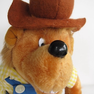 May include: A brown plush bear wearing a brown cowboy hat and blue overalls. The bear has a yellow and white checkered shirt and a button on the overalls. The bear has a black nose and white eyes.
