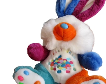 May include: A colourful plush bunny toy with a white body, blue and purple ears, and orange, blue, and green accents. The bunny has a white face with a large orange nose and blue eyes. The bunny is wearing a white shirt with a colourful jelly bean design and the text "Jelly Bean" on the front.