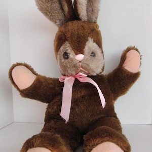 Puede incluir: Un conejo de peluche marrón con orejas rosas y una cinta rosa alrededor del cuello. El conejo está sentado con los brazos extendidos.