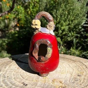 Puede incluir: Una pequeña casita de pájaros de cerámica roja con un agujero para que los pájaros entren. La casita tiene un diseño marrón y verde alrededor del agujero y una flor amarilla en la parte superior.