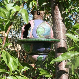 Peut inclure: Un nichoir à oiseaux en céramique avec un motif vert, rose et violet suspendu à une branche d'arbre. Le nichoir a un dessus rond ouvert et une plaque inférieure.