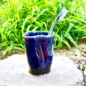 May include: A handmade ceramic toothbrush holder with a deep blue glaze and a toothbrush. The holder has a unique shape and a glossy finish. The toothbrush has a blue and yellow handle and white bristles.
