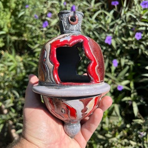 Peut inclure: Une décoration en céramique rouge, blanche et grise avec un trou au centre. La décoration a une surface texturée et un petit bouton gris sur le dessus.