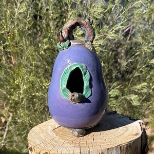 Puede incluir: Una casita de pájaros de cerámica morada con forma de pera, con un detalle de hoja verde y un pequeño agujero para que los pájaros entren. La casita de pájaros está sentada sobre un tocón de árbol.