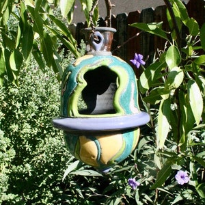 Peut inclure: Un nichoir à oiseaux en céramique en forme de gourde avec un glaçage vert, bleu et jaune. Le nichoir est suspendu à une branche d'arbre.