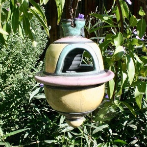 Peut inclure: Un nichoir à oiseaux en céramique avec un glaçage jaune, rose et vert. Le nichoir a une forme ronde avec une petite ouverture pour que les oiseaux puissent entrer. Il est suspendu à une branche d'arbre.