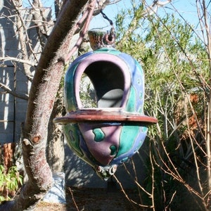 Peut inclure: Un mangeoire à oiseaux en céramique colorée avec un motif rose, vert et bleu. La mangeoire est suspendue à une branche d'arbre.