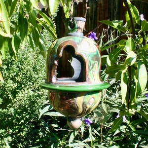 Peut inclure: Un nichoir à oiseaux en céramique vert, brun et jaune avec un design décoratif suspendu à une branche d'arbre. Le nichoir a une grande ouverture pour que les oiseaux puissent accéder à la nourriture.