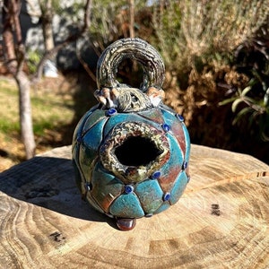 Puede incluir: Un comedero para pájaros de cerámica turquesa y marrón hecho a mano con un asa circular texturizada. El comedero para pájaros presenta una abertura redonda y está adornado con detalles azules y marrones. Se exhibe sobre una superficie de madera natural.