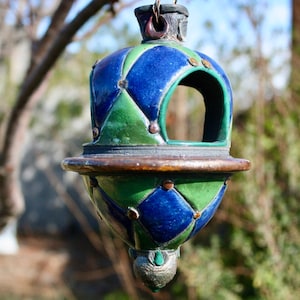 Peut inclure: Un nichoir à oiseaux en céramique bleu et vert avec un motif en losange décoratif et une petite ouverture pour que les oiseaux puissent entrer. Le nichoir est suspendu à une chaîne.