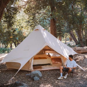May include: A large, beige canvas tent pitched in a forest setting. The tent has a bell shape with a central pole and is open at the front, revealing a bed inside. A person sits in a chair outside the tent.