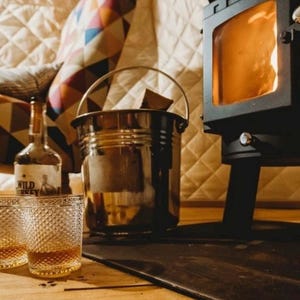 May include: A cozy scene featuring a small black stove with a glowing fire, a metal bucket, two glasses, and a bottle of Wild Turkey whiskey. The glasses and bottle are on a wooden surface, suggesting a warm and inviting atmosphere.