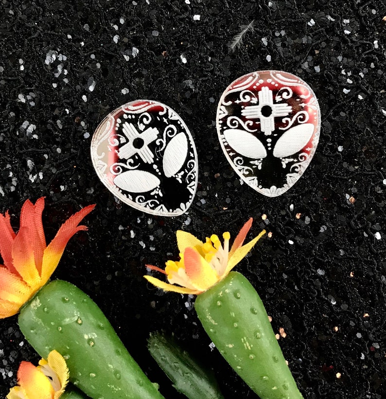 May include: Two white alien face earrings with black and white details on a black background. The earrings are made of resin and have a glossy finish.