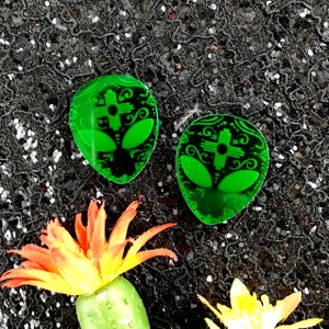 May include: Two green alien-shaped earrings with a cross-like design in the center. The earrings are on a black sparkly background.