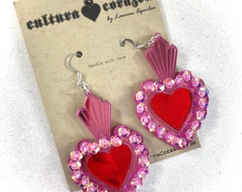 May include: Pink and red heart-shaped earrings with a mirror in the center and pink sequins around the edges. The earrings are on a card that says "cultura corazón by Lauren Aguilar".