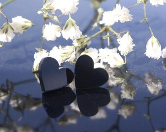 May include: Two black heart-shaped earrings on a reflective surface with white flowers in the background.