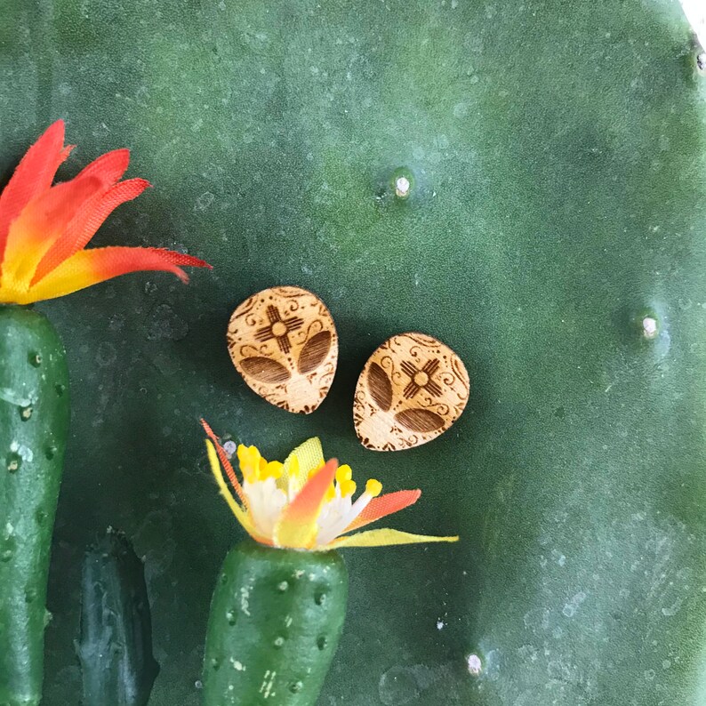 May include: Two wooden earrings with a geometric design. The earrings are light brown and have a pattern of a cross with four points. The earrings are on a green cactus background.