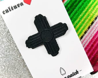 May include: Black iron-on patch featuring a stylized black cross with a circle in the center. The patch is on a white card with the text "cultura corazon" and "iron on patch - culturaCorazon.com".