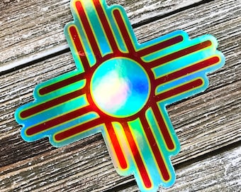 May include: A holographic sticker featuring the Zia symbol, a traditional symbol of the Pueblo people of New Mexico. The symbol is a red and yellow sun with four rays extending from it.