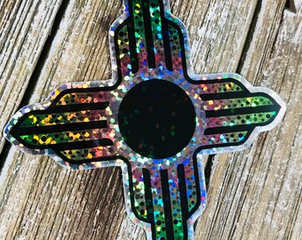 May include: A holographic sticker of the Zia symbol, a traditional symbol of the Pueblo people of New Mexico. The symbol is a stylized sun with four rays, each with a series of dots. The sticker is iridescent and has a black center.