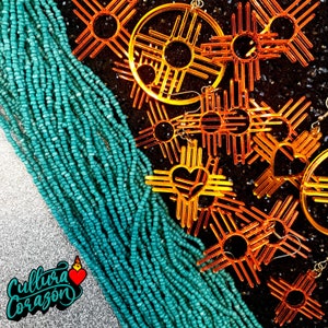 Peut inclure: Un collier de perles turquoise avec plusieurs boucles d'oreilles en forme de soleil orange et brun. Les boucles d'oreilles ont la forme d'un soleil à quatre pointes et un cercle au centre. Les boucles d'oreilles sont superposées les unes sur les autres.