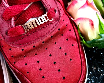 May include: A close-up of a red sneaker with a gold chain that says "Amore" on it. The sneaker is surrounded by a white rose and green leaves.