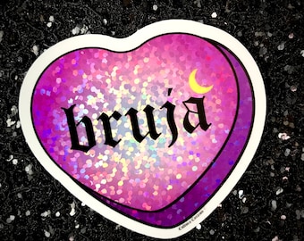 May include: A pink heart-shaped sticker with a holographic glitter finish. The sticker features the word "bruja" in black gothic lettering and a crescent moon in yellow.