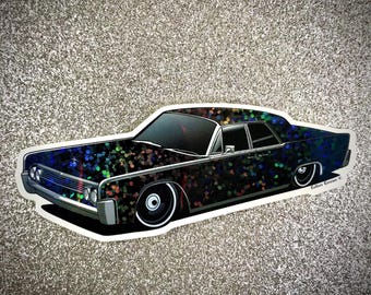 May include: A sticker featuring a classic black car with a lowrider design. The car has a dark, speckled pattern with iridescent accents. The sticker is outlined in white, and the background is a glittery silver.