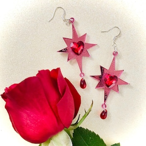 May include: A pair of pink star-shaped earrings with a red heart-shaped crystal in the center of each star. The earrings have a dangling red crystal drop.