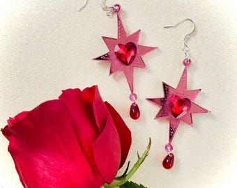 May include: A pair of pink star-shaped earrings with a red heart-shaped crystal in the center of each star. The earrings have a dangling red crystal drop.