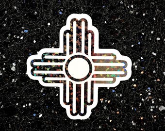 May include: A white sticker with a black outline of the Zia symbol, a traditional symbol of the Pueblo people of New Mexico. The symbol is filled with iridescent glitter.