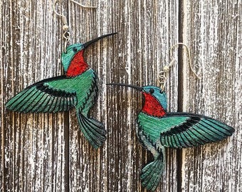 May include: A pair of hummingbird earrings with a teal blue and green body, red throat, and black wings. The earrings are made of wood and have silver hooks.