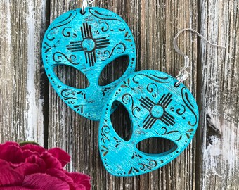 May include: Teal blue alien face earrings with a black and white southwestern design. The earrings are made of wood and have a distressed finish.