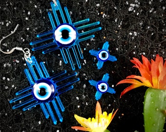 May include: A pair of earrings with a blue evil eye design in the center of a cross shape. The earrings are made of metal and have a silver finish. The evil eye is a symbol of protection against bad luck and envy.