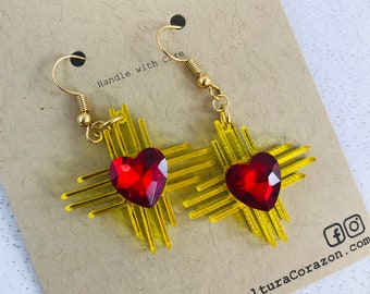 May include: A pair of gold earrings with a red heart and yellow geometric design. The earrings are on a brown card with the text "Handle with Care" and "CulturaCorazon.com".