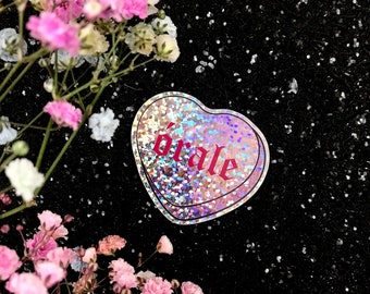 May include: A holographic heart-shaped sticker with the word "orale" written in red glitter. The sticker is on a black background with silver glitter.