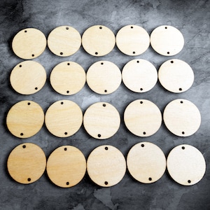 Peut inclure: Vingt et un cercles en bois non finis avec un grain de bois naturel. Chaque cercle a un petit trou en haut et deux petits trous en bas. Les cercles sont disposés en rangées et en colonnes sur un fond gris foncé.