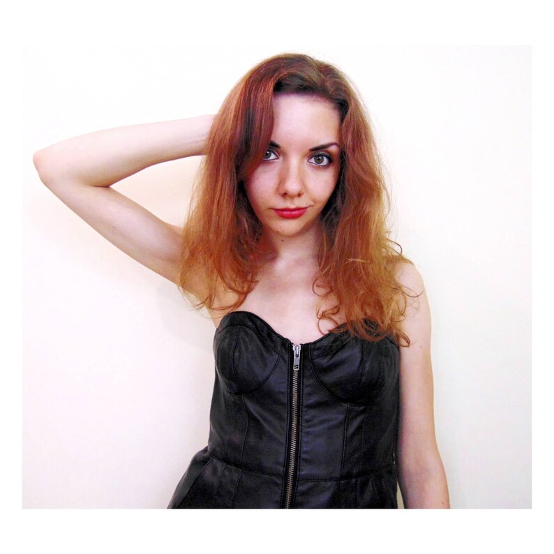 May include: A woman with long, wavy, auburn hair is wearing a black strapless dress with a zipper down the front. She is looking at the camera with a serious expression.