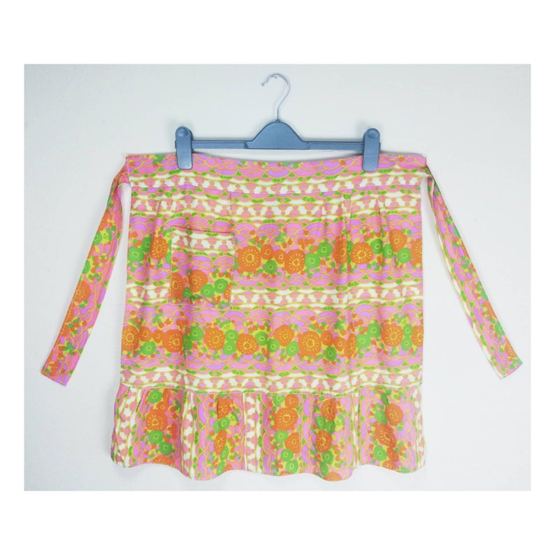May include: A pink and green floral print apron with a ruffled hem and two long ties. The apron has a single pocket on the front.