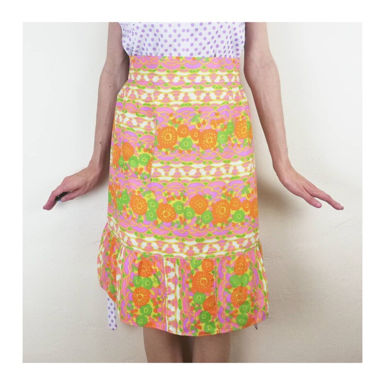 May include: A pink and green floral print apron with a ruffled hem and a large pocket. The apron has a retro 1960s style.