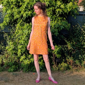 Peut inclure: Une robe courte sans manches avec un motif chevron orange et rose vif, un col montant et un cordon de serrage jaune à la taille. La robe présente une coupe droite arrivant au-dessus du genou, portée avec des chaussures plates rose vif.