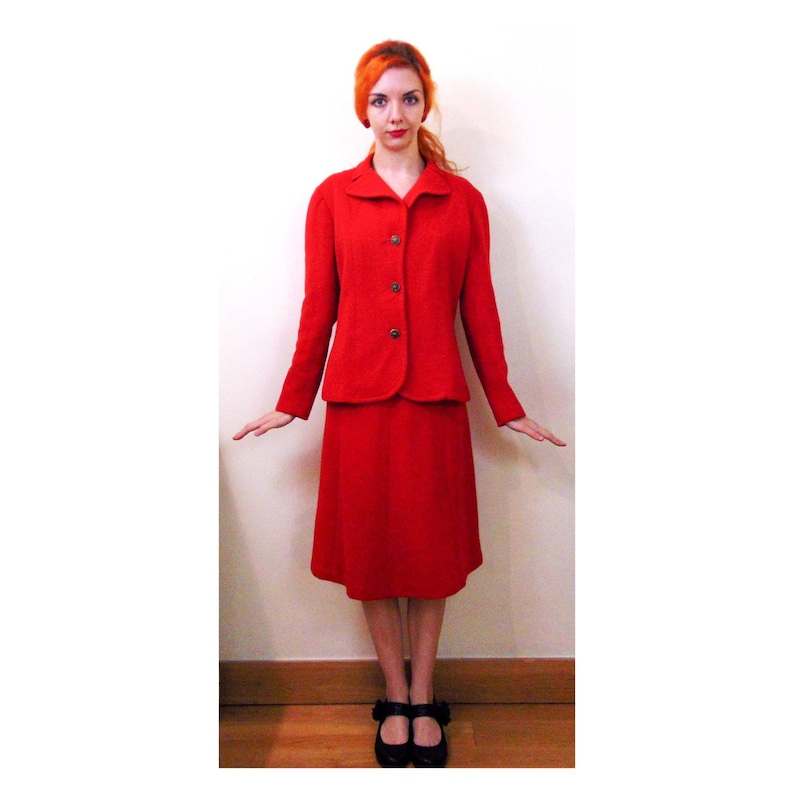 May include: A red wool suit with a skirt and a jacket. The jacket has a button closure and a collar. The skirt is knee-length.
