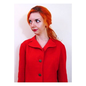 May include: A woman with bright orange hair is wearing a red wool jacket with a collar and four buttons. The jacket has a simple design and is a classic style.
