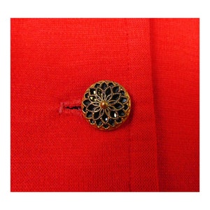 May include: A close-up of a gold-coloured button with a black centre and a decorative design. The button is on a red fabric background.
